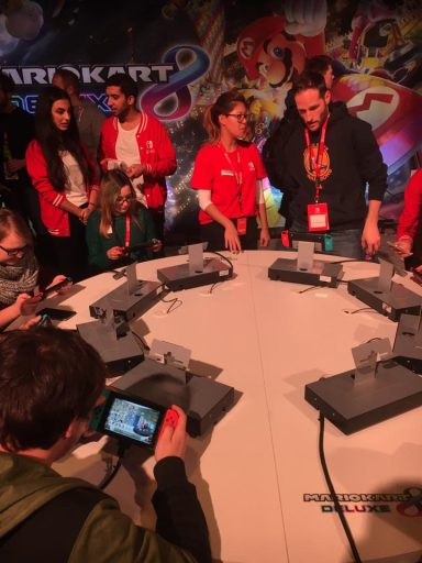 Mehrere Personen in roten T-Shirts spielen an Nintendo Switch Konsolen an einem Tisch.