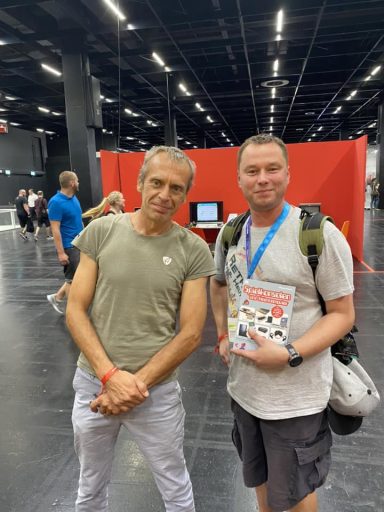 Zwei Männer stehen in einer Messehalle, einer hält ein Buch in der Hand.