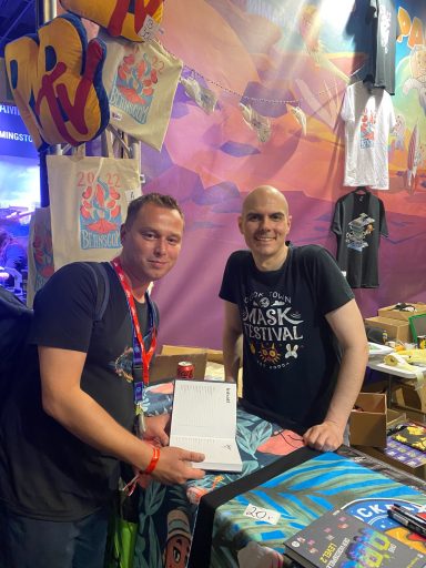 Zwei Männer stehen an einem Veranstaltungstisch mit Merchandise-Artikeln.